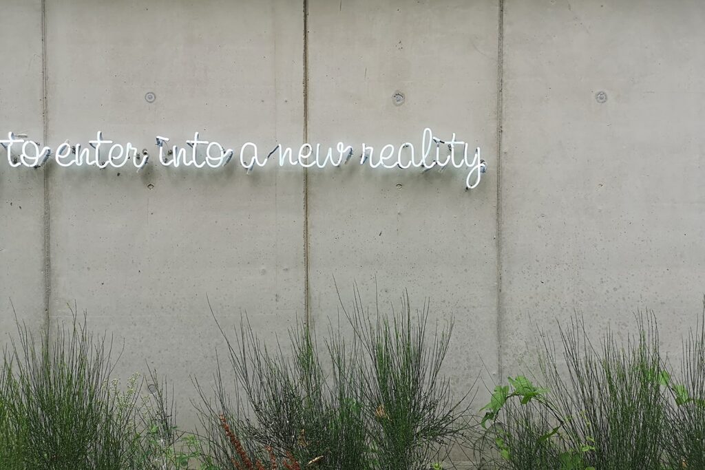 Weiße Neonschrift vor Betonwand mit Gras davor